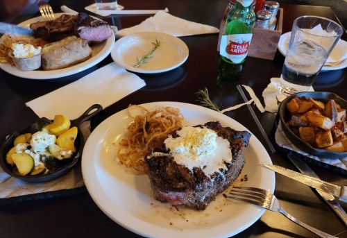 A plate of steak topped with butter, served with crispy onions and sides of potatoes and vegetables, with drinks on the table.