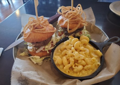 Two mini burgers topped with crispy onions on a plate, accompanied by a small skillet of macaroni and cheese.