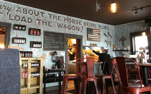 Interior of a rustic restaurant with wooden walls, a chalkboard menu, and red metal chairs. A person is at the counter.