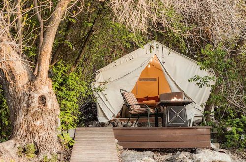 A cozy glamping tent on a wooden deck, surrounded by greenery, with a fire pit and seating area outside.