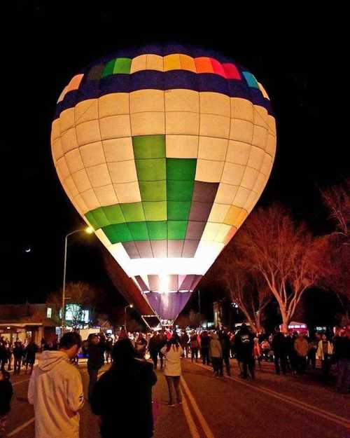 A brightly lit hot air balloon rises at night, surrounded by a crowd on a street. Trees and buildings are in the background.