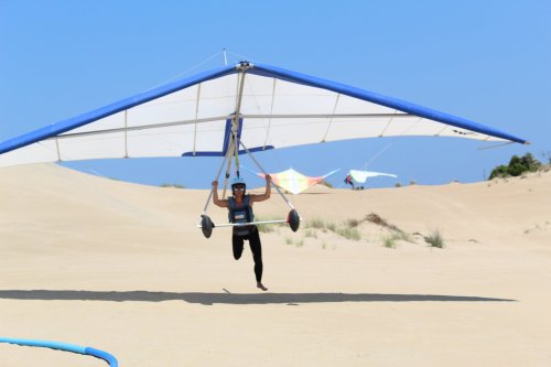 A person hang gliding over sandy dunes under a clear blue sky, preparing for takeoff.