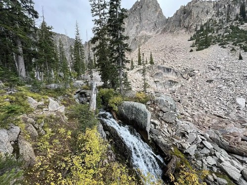 A serene mountain landscape featuring a small waterfall, rocky terrain, and evergreen trees under a cloudy sky.