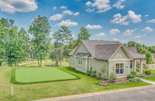 A modern house beside a green lawn with a putting green, surrounded by trees and a blue sky with fluffy clouds.
