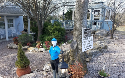 A blue-clad mannequin stands by a tree, with a sign for check-in and a gift shop in a landscaped outdoor area.
