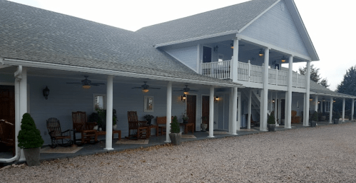 A light blue building with rocking chairs on a porch, surrounded by gravel and greenery.