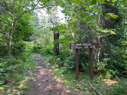 A wooded path with a sign for "The Grand Portage" indicating distances to various locations. Lush greenery surrounds the trail.