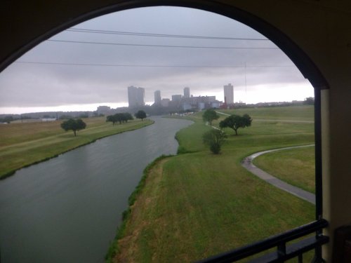 A view of a river winding through green fields, with a city skyline visible in the background under a cloudy sky.