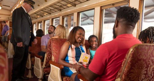 A family enjoys snacks on a vintage train, smiling and chatting with a conductor in the background.