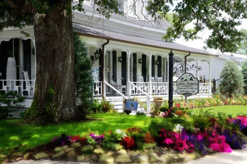 A charming white house with a porch, surrounded by colorful flowers and greenery, featuring a sign for a bed and breakfast.
