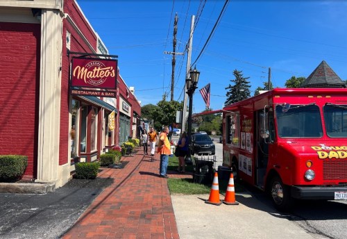 A red food truck parked beside a restaurant on a sunny day, with people walking on the sidewalk and an American flag nearby.