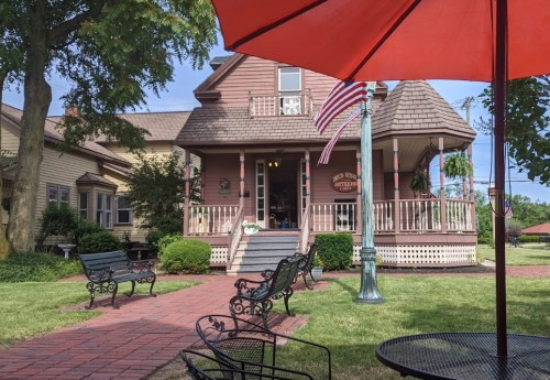 A charming two-story house with a porch, surrounded by greenery and benches, under a bright red umbrella.