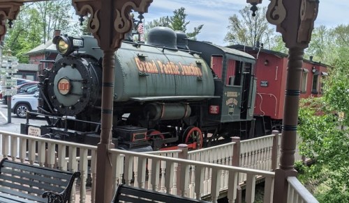 A vintage steam locomotive and red caboose displayed near a gazebo, surrounded by greenery and parked cars.