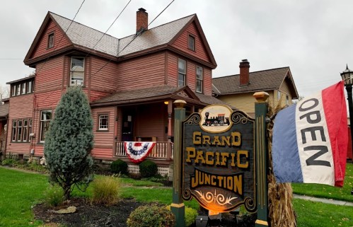 A historic red house with a sign reading "Grand Pacific Junction" and an open flag, surrounded by greenery.