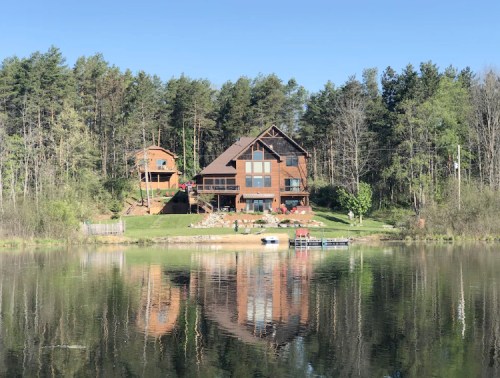 A large wooden house by a calm lake, surrounded by trees, with a smaller building nearby and a dock in the water.