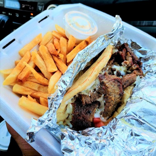 A hand holding a takeout container with fries and a wrap filled with meat and vegetables, accompanied by a small sauce cup.