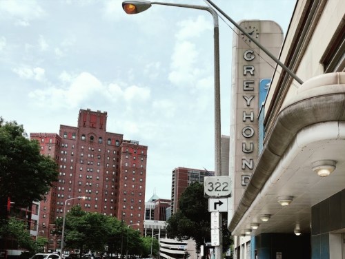 View of a Greyhound bus station with tall buildings and a street sign in the foreground under a cloudy sky.