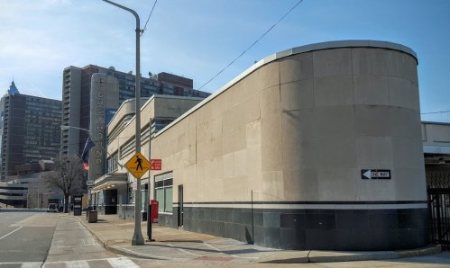 Exterior view of a Greyhound bus station with a modernist design, surrounded by urban buildings and a clear blue sky.