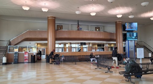 Interior of a bus station with benches, a ticket counter, and a large screen, featuring a few waiting passengers.