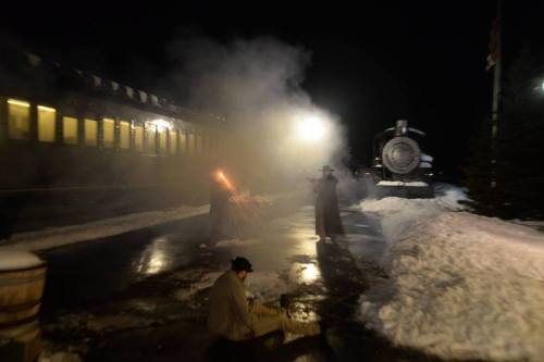 A snowy train station at night, with smoke and two figures near a fire, and a steam locomotive in the background.