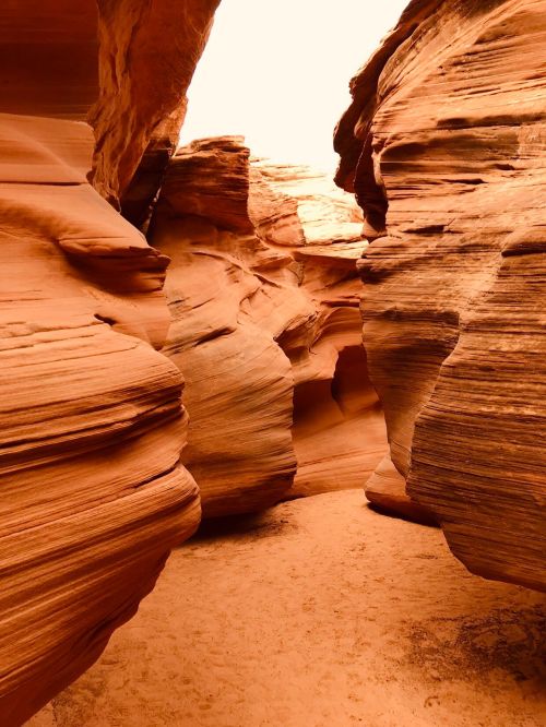 A narrow canyon with smooth, layered red rock formations and soft sand under a bright sky.