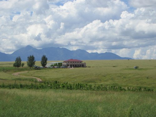 A spacious green field with a house in the distance, surrounded by mountains and a cloudy sky.