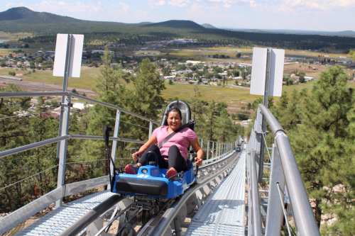 A woman rides a mountain coaster down a track with scenic hills and a town in the background.