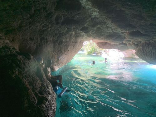 A cave opening reveals turquoise water and people swimming inside, with rocky walls and greenery in the background.