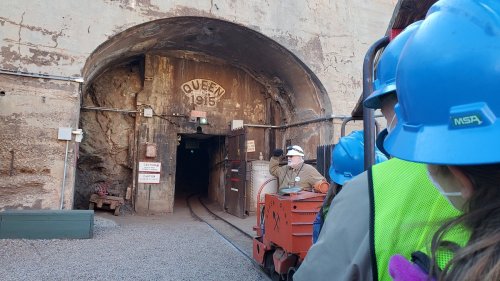A mine entrance labeled "Queen 1915," with a guide and visitors wearing hard hats, ready to explore.