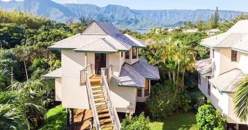 A two-story house with a staircase, surrounded by lush greenery and mountains in the background.