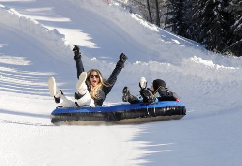 A person joyfully tubing down a snowy slope, arms raised, with another person behind them on a tube.