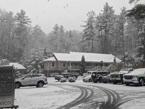 A snowy scene with several cars parked in a lot, surrounded by trees and a building covered in snow.