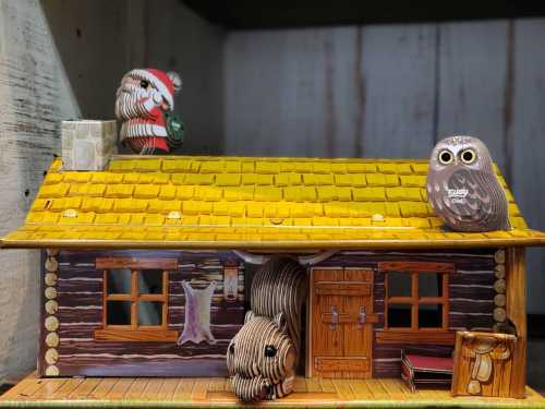 A whimsical wooden cabin with a yellow roof, featuring two owls perched on top and a wooden bear in front.