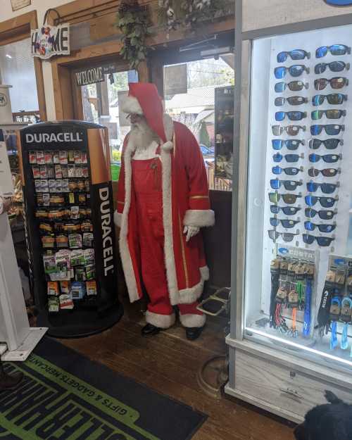 A life-sized Santa Claus figure stands inside a store next to a display of sunglasses and batteries.