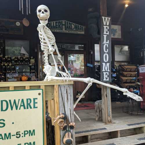 A skeleton sits on a wooden railing outside a hardware store, with a "Welcome" sign and store hours visible.