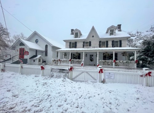 A snowy scene featuring a charming white house with a porch, decorated with red bows, and a nearby church.