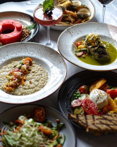 A variety of beautifully plated dishes, including seafood, salads, and a colorful dessert, with drinks in the background.