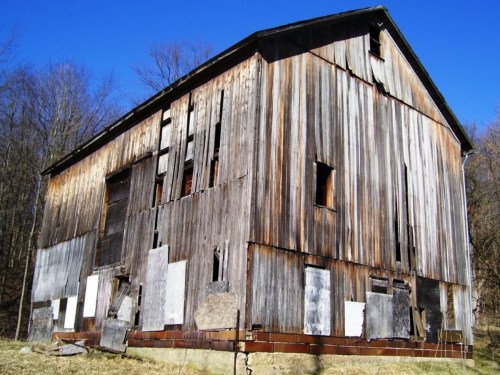 An old, weathered barn with peeling wood and patches of metal, set against a clear blue sky and trees in the background.