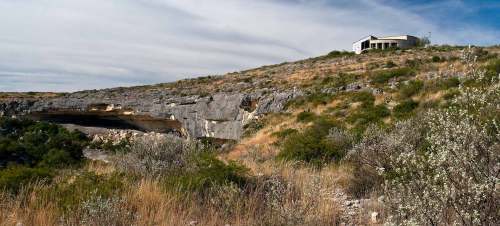 A hillside with sparse vegetation and a house perched on top, overlooking rocky terrain and a cloudy sky.