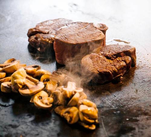 Sizzling steaks and sliced mushrooms on a hot grill, with steam rising in the air.