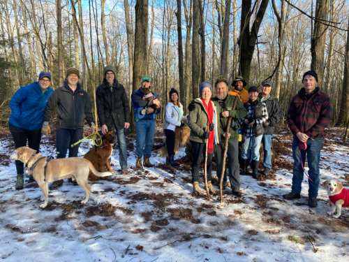A group of people and dogs stand together in a snowy forest, smiling and enjoying the outdoors.