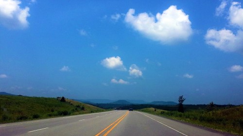 A wide, empty road stretches into the distance under a bright blue sky with fluffy white clouds and green hills on either side.