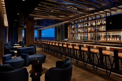 A modern bar with blue seating, a well-stocked shelf of spirits, and large windows showcasing a nighttime view.