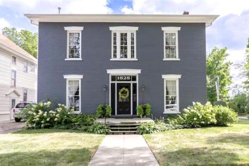 A large gray two-story house with white trim, a front porch, and lush greenery in the yard.