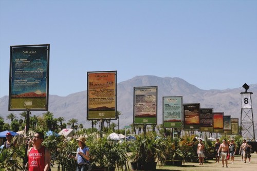 Festival-goers walk past large posters displaying Coachella lineups from 2007 to 2008, with mountains in the background.