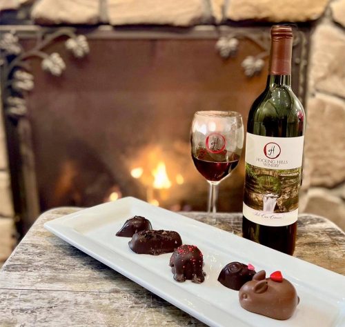 A plate of chocolate treats beside a glass of red wine and a bottle, with a cozy fireplace in the background.