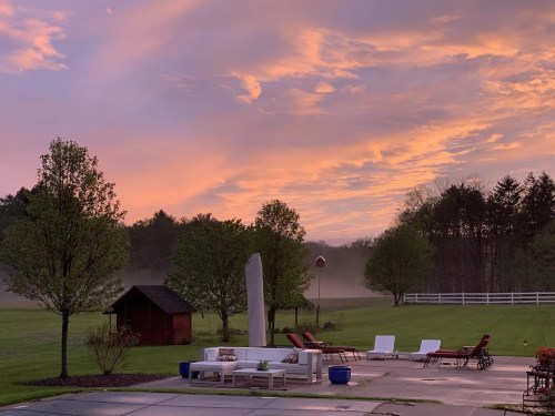 A serene landscape at sunset, featuring a misty field, trees, and lounge chairs by a patio.