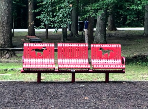 Three red park benches with dog silhouettes, set in a wooded area with trees in the background.