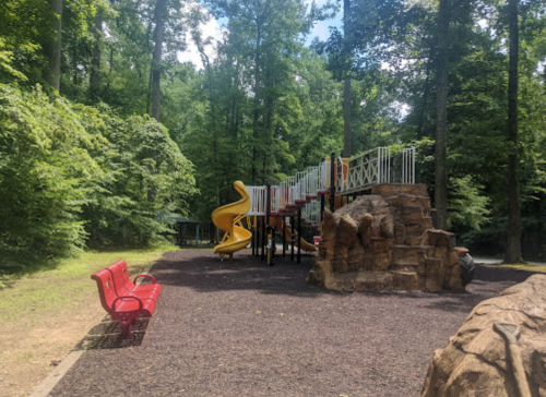 A playground with a yellow slide and climbing structure, surrounded by trees, with a red bench in the foreground.