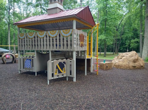 A wooden playground structure with a slide, surrounded by trees and a rocky area, on a rubberized surface.